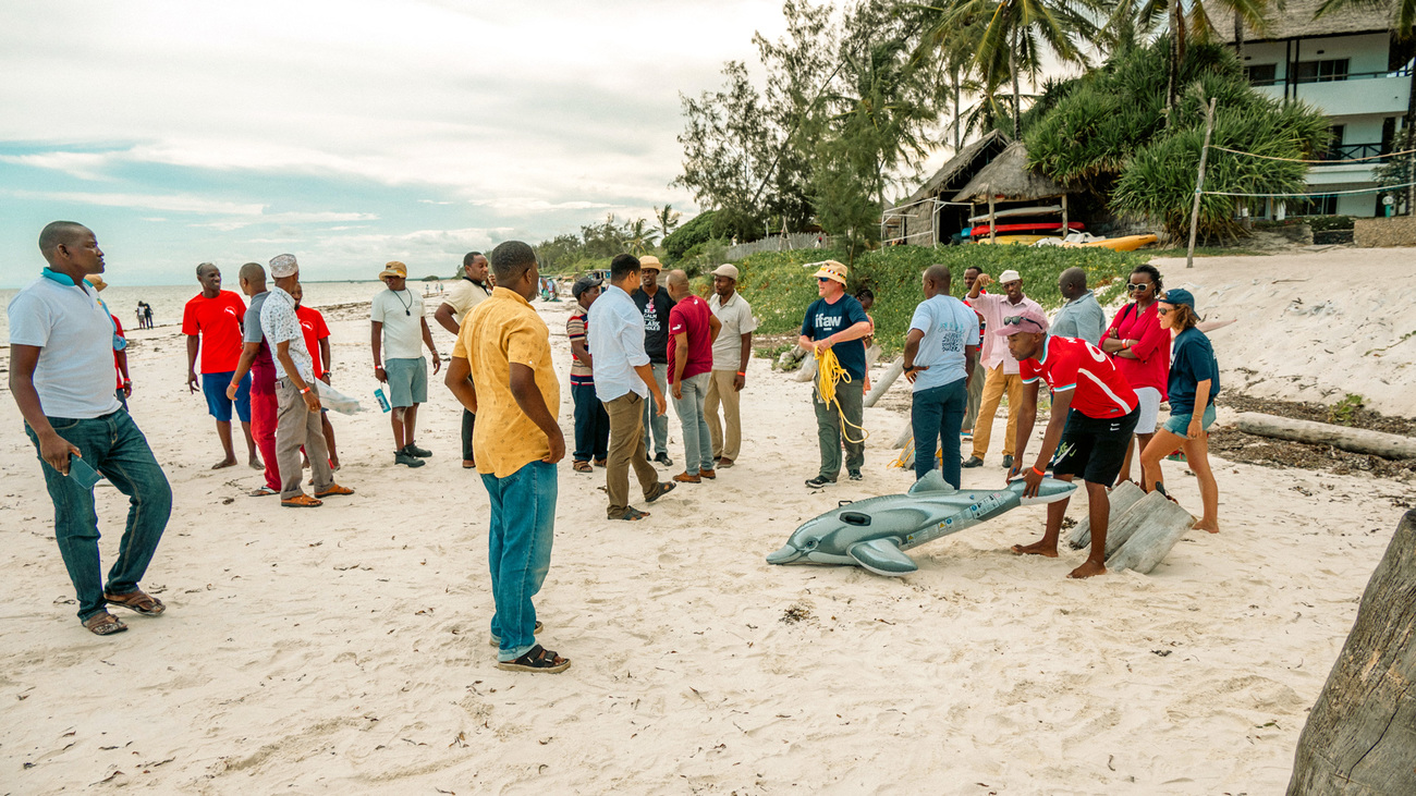 Viele Menschen stehen zusammen an einem Strand. Ein Delfinimitat liegt auf dem Boden.