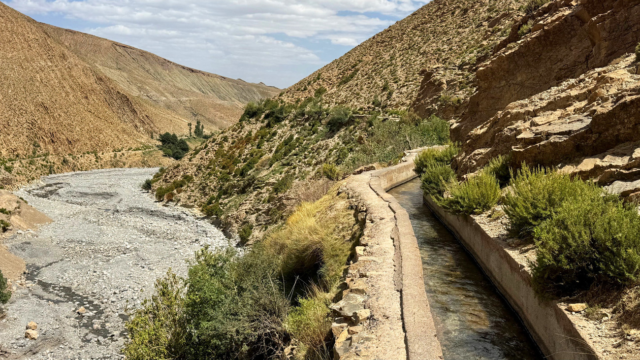 A picturesque irrigation canal winds through a dry valley in Morocco, surrounded by rugged hills under a cloudy sky.