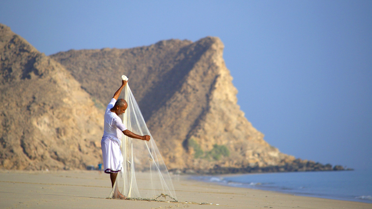 A fisherman casts his net on the serene As Sifah beach by the rocky Omani coastline.